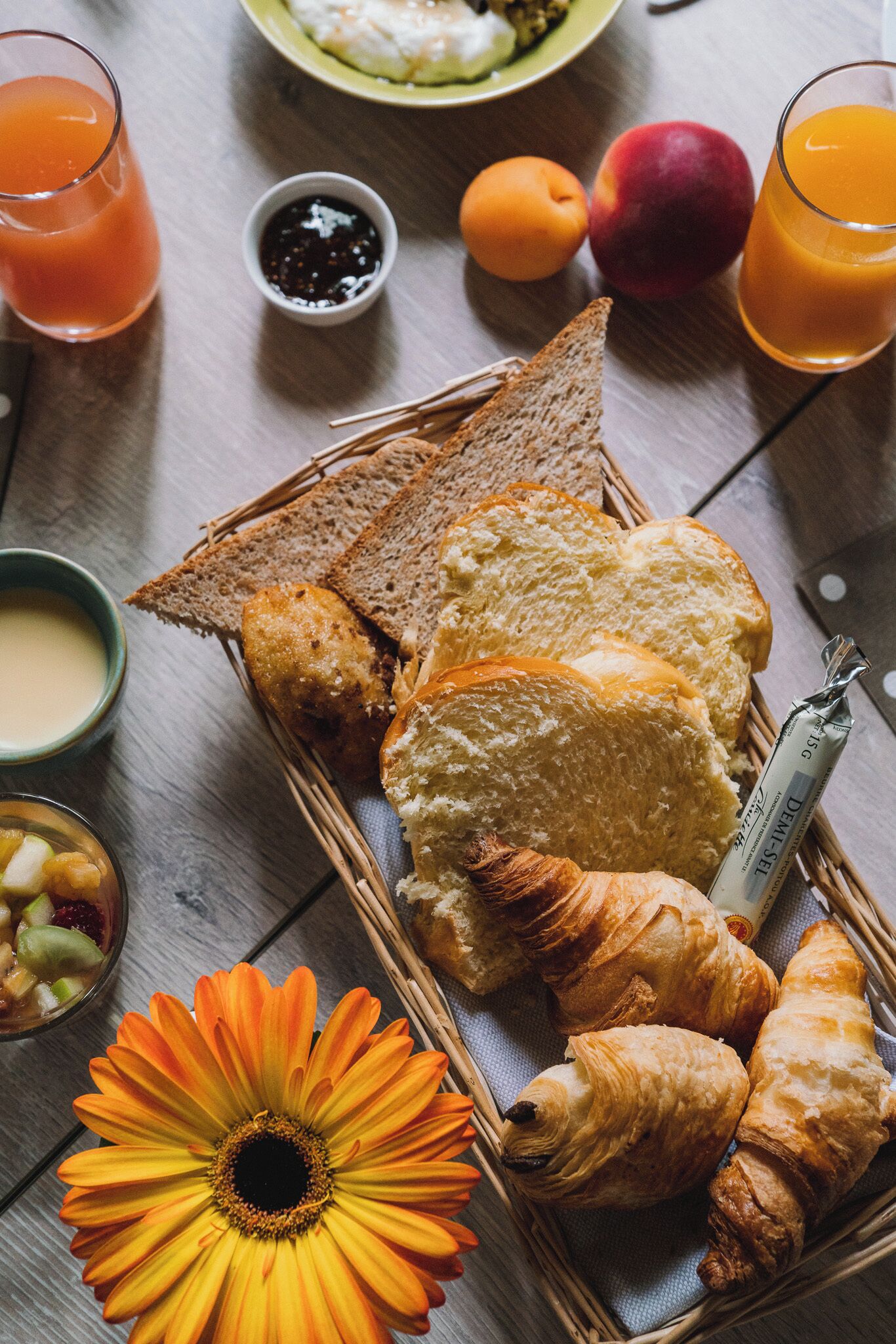 A breakfast setting with bread, croissants, a flower, and drinks on a wooden table.