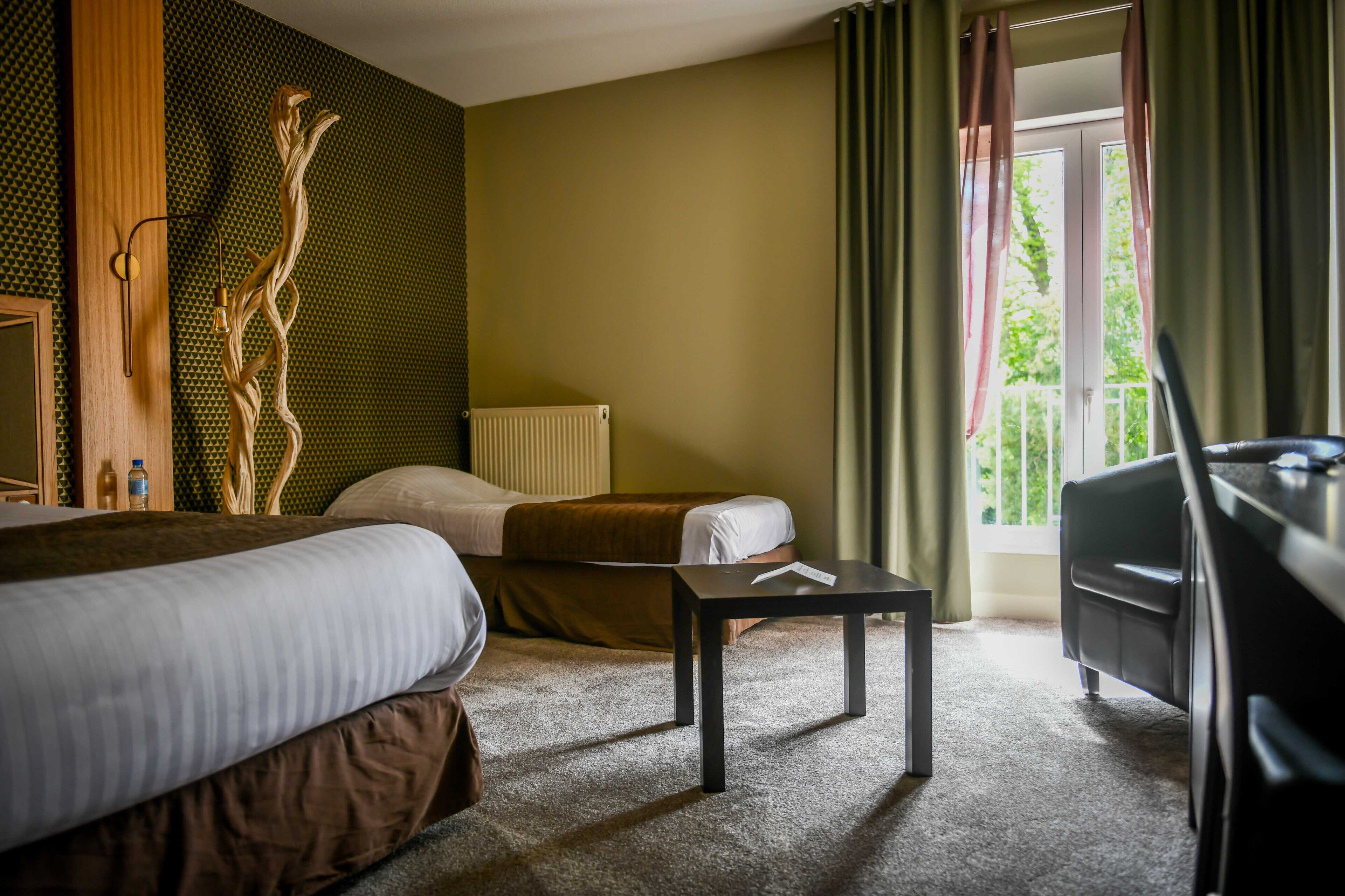 Une chambre d’hôtel avec deux lits, une petite table, un radiateur et une vue sur la verdure par la fenêtre.