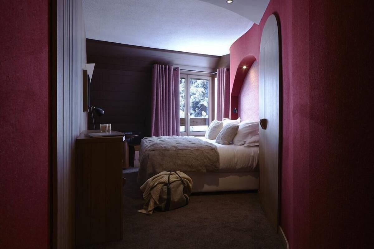 Un dormitorio con una cama, un escritorio, una silla, una ventana con cortinas y una bolsa en el suelo.