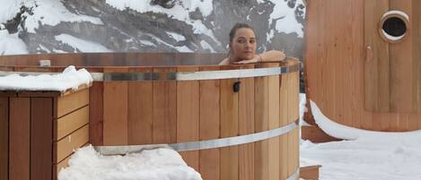 Una bañera de madera rodeada de árboles cubiertos de nieve y un edificio al fondo.