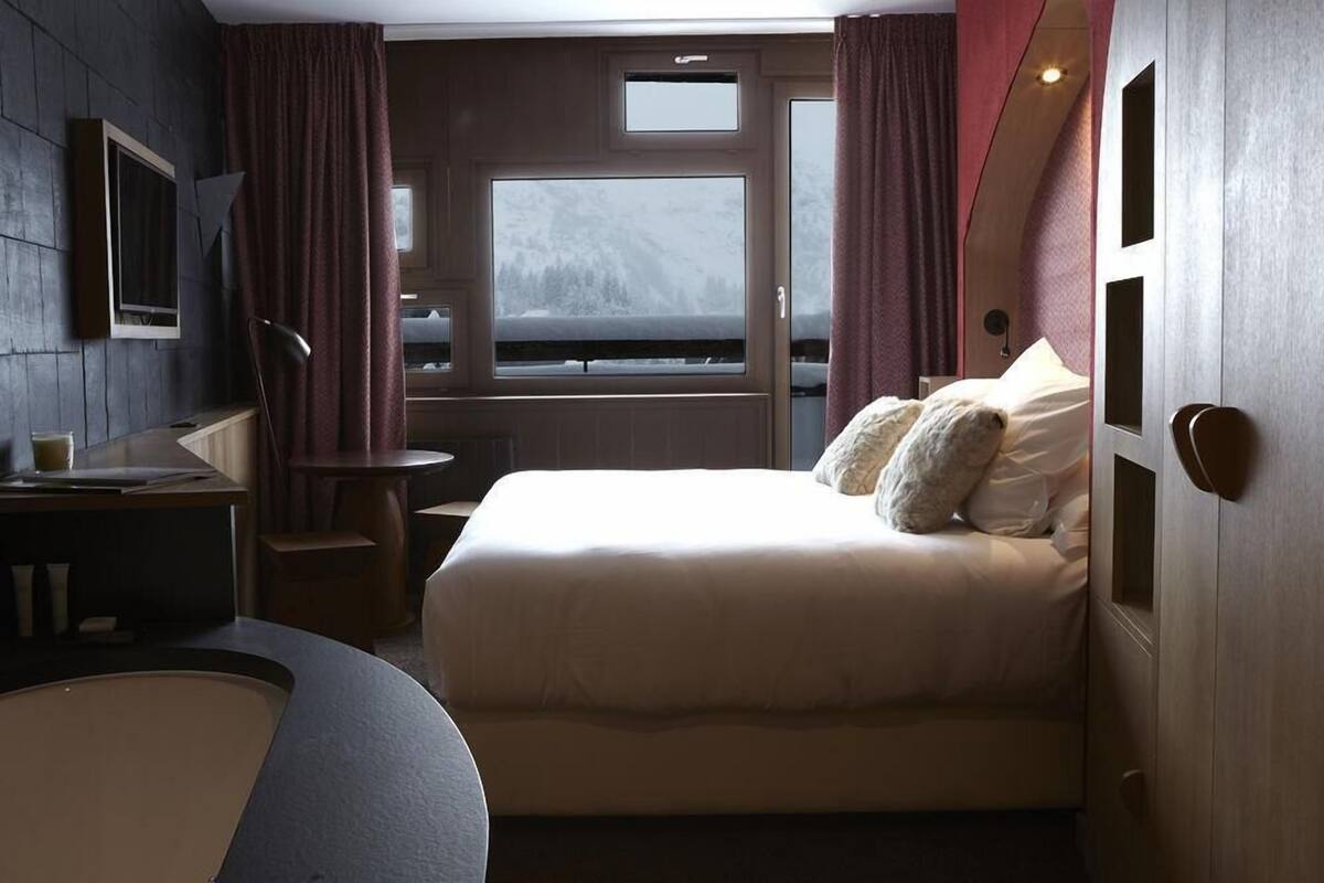 Un dormitorio con cama, escritorio, silla, ventana y vista a un paisaje nevado, además de una puerta.