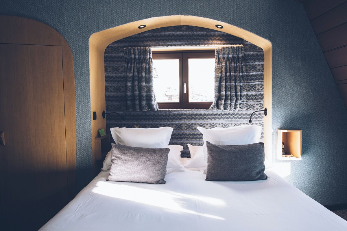 Une chambre à coucher avec un lit, deux oreillers, une fenêtre avec des rideaux, une porte en bois et une petite étagère sur laquelle se trouve une bouteille.