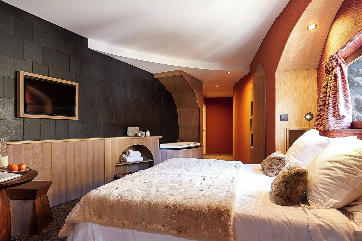 Une chambre d’hôtel moderne avec un mur en pierre sombre, une coiffeuse en bois, un grand lit avec une couette moelleuse et une ambiance chaleureuse.