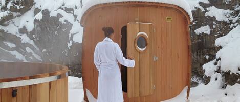 Una estructura de sauna de madera con puerta circular, rodeada de árboles cubiertos de nieve y un edificio al fondo.