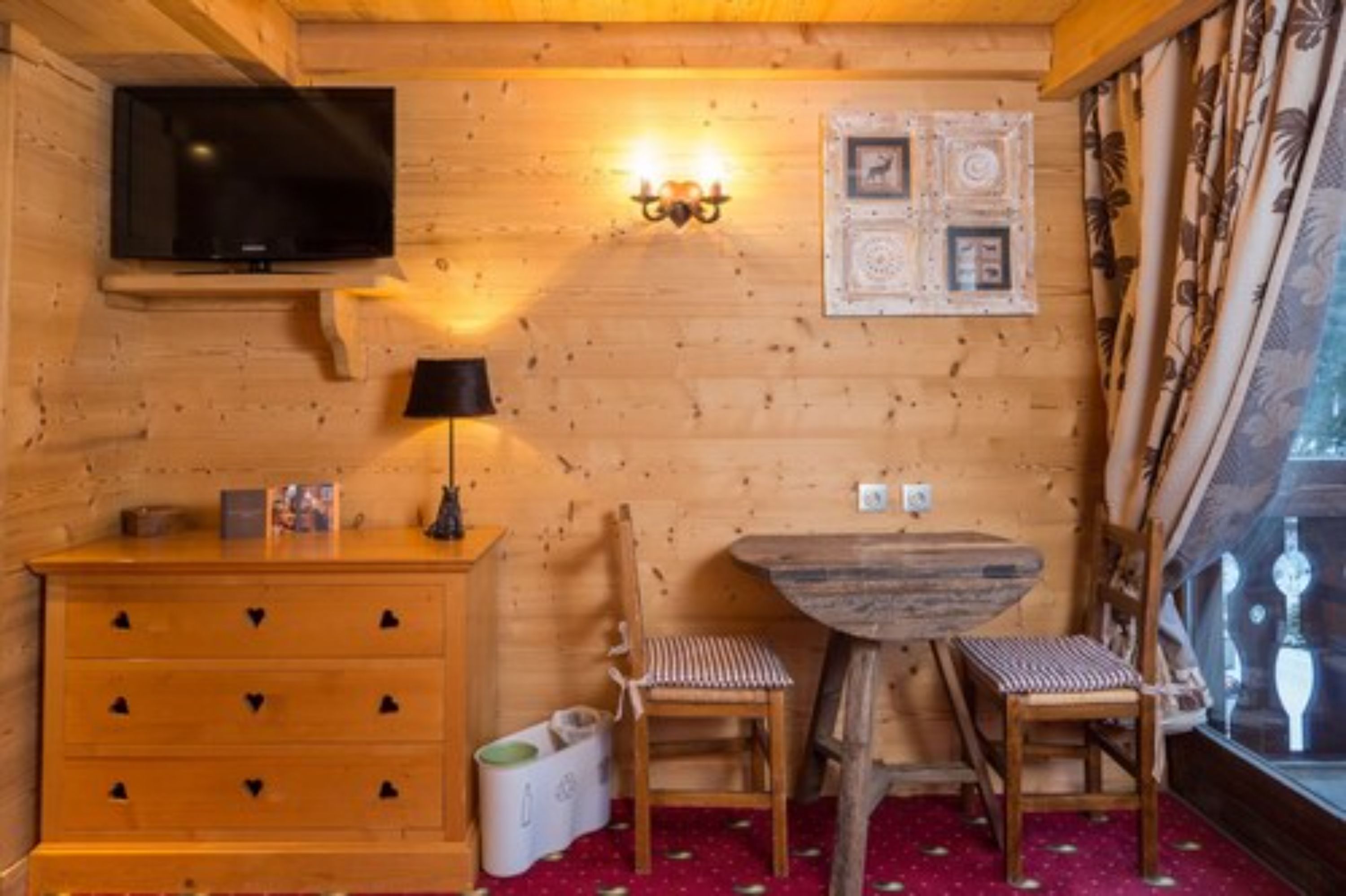 A cozy wooden cabin room with a TV, a wall-mounted light, a wooden dresser, two chairs, and a small table.
