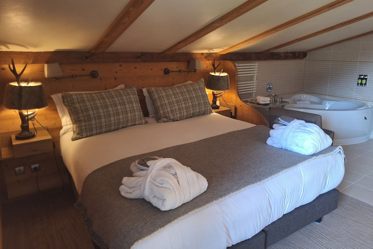 Ein Schlafzimmer mit einem großen Bett, zwei Nachttischen, einer Lampe sowie einem Badezimmer mit einer Badewanne und Holzbalken an der Decke.