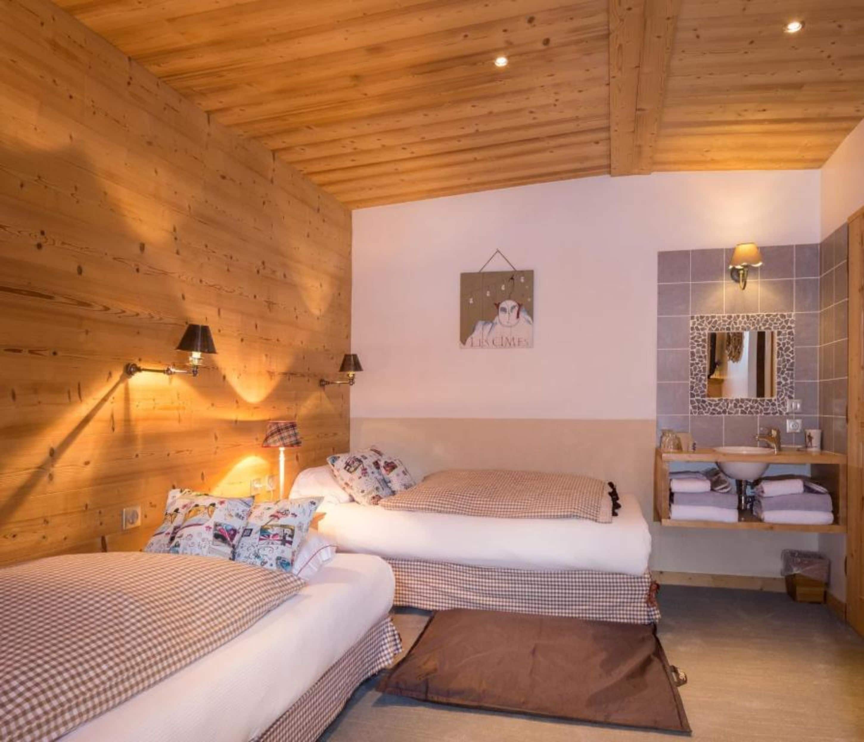 A cozy room with wooden walls and ceiling, two beds with checkered bedding, a wall-mounted mirror, and a small bathroom with a sink and towel rack.