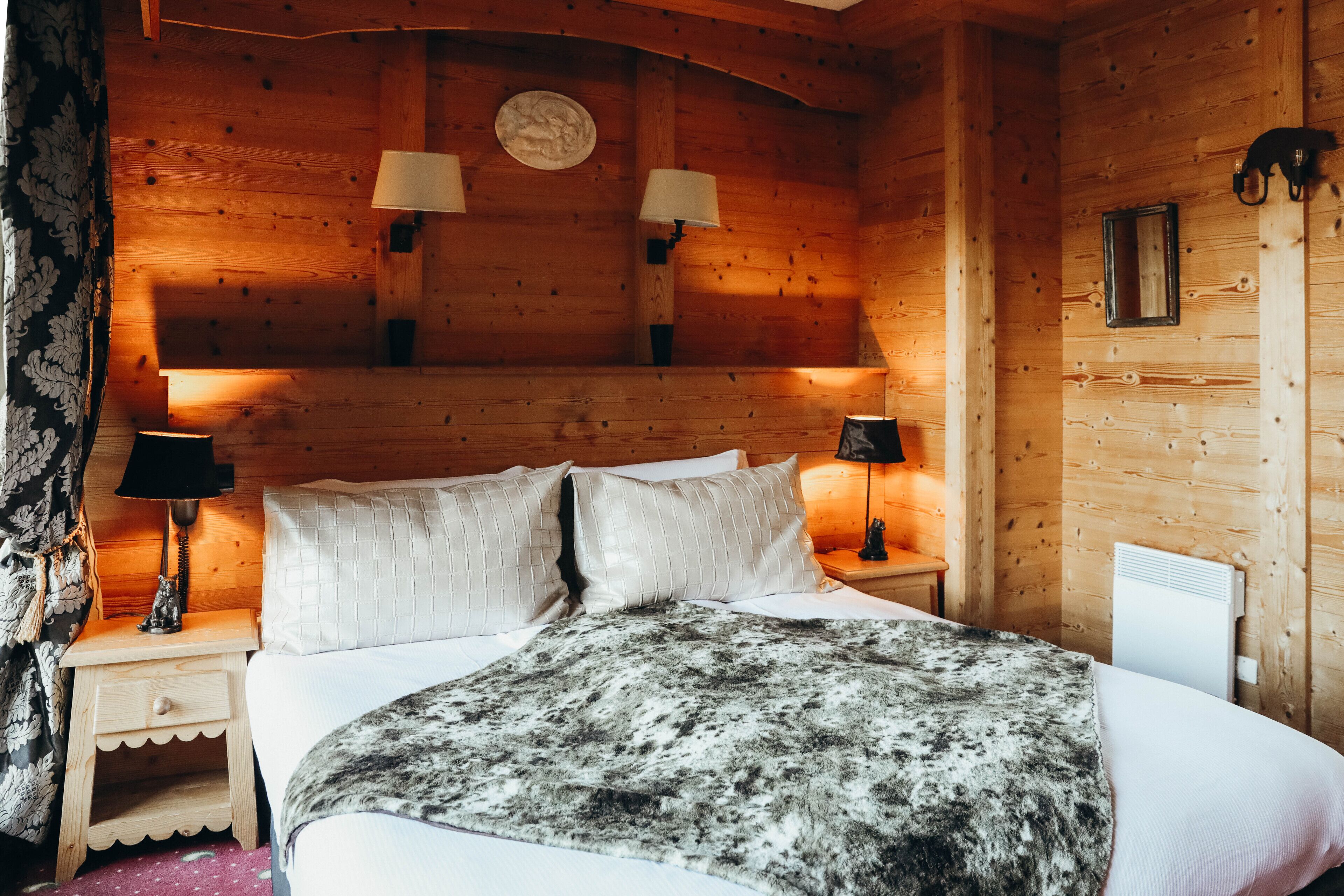 A wooden cabin-style bedroom with a bed, two bedside lamps, a nightstand, and a wall-mounted heater.