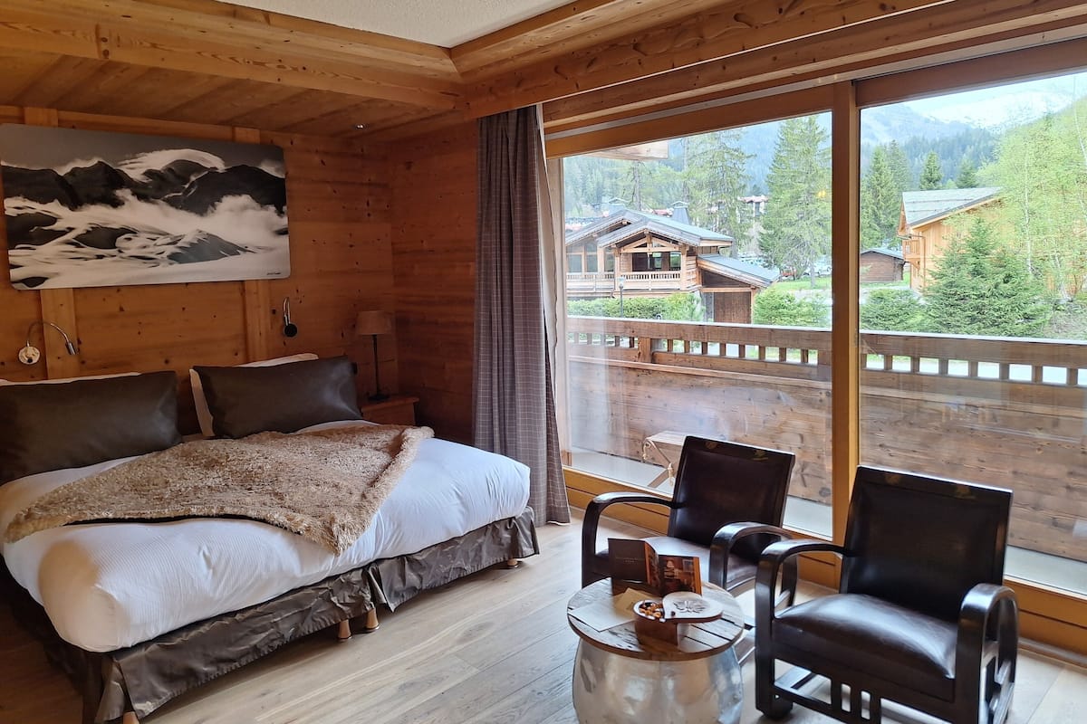 Ein Schlafzimmer einer Holzhütte mit einem Bett, zwei Stühlen, einem kleinen Tisch und einem großen Fenster mit Blick auf einen Berg und einen Wald.