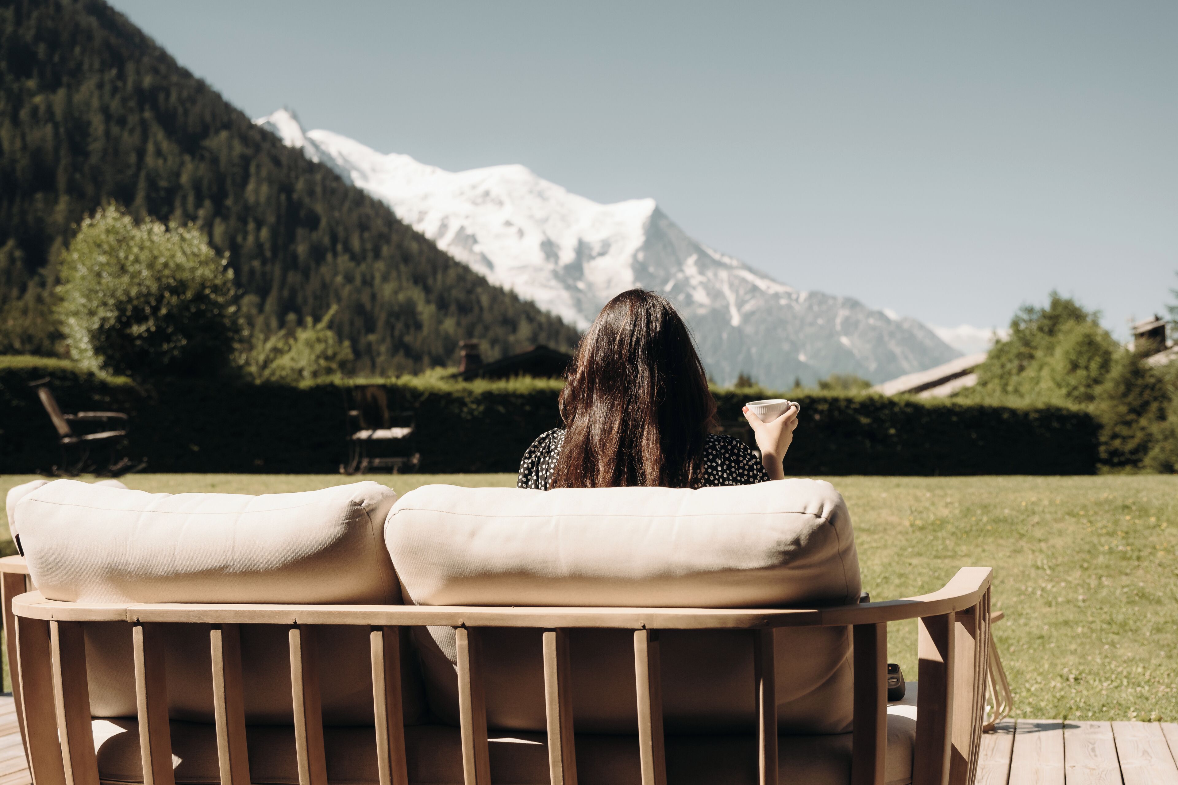 A person sitting on a wooden bench with cushions, facing a snow-capped mountain.