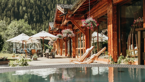 A wooden cabin with a pool, outdoor seating, and hanging flower baskets.