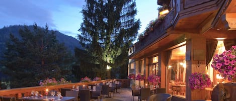 An outdoor dining area with tables and chairs, surrounded by flowers and a large tree.
