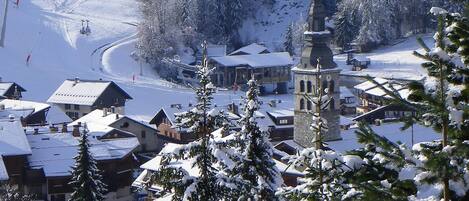 Un pueblo cubierto de nieve con edificios tradicionales, una iglesia con una torre destacada y una pista de esquí al fondo.