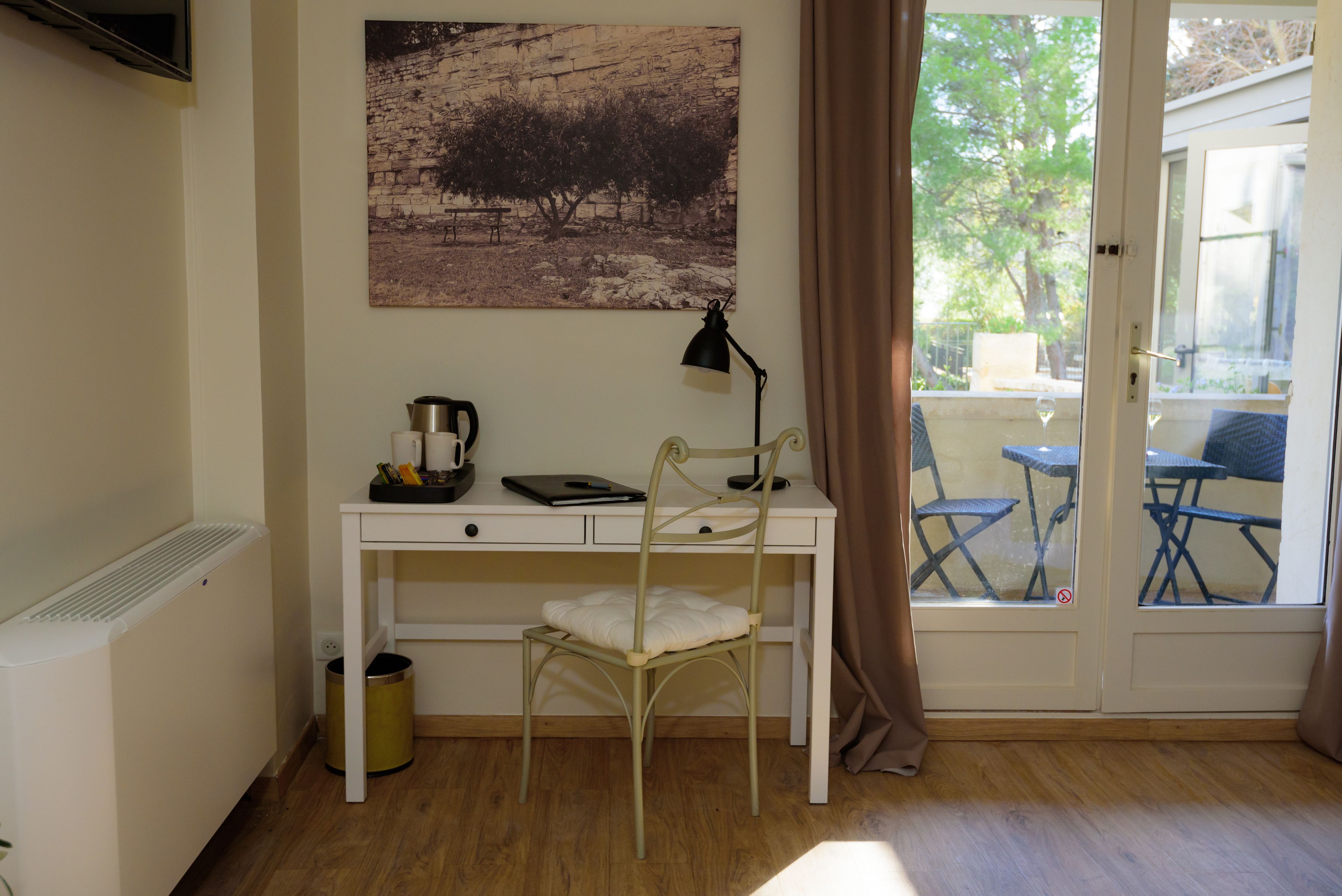 A room with a white desk, a chair, a radiator, a picture on the wall, and a view of outdoor furniture through a glass door.