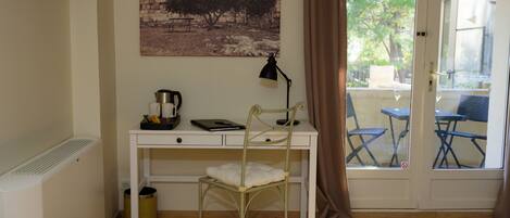 A room with a white desk, a chair, a radiator, a picture on the wall, and a view of outdoor furniture through a glass door.