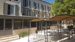 A two-story building with a balcony, outdoor seating area with tables and chairs, and a large tree in the background. - Hôtel Belesso (Fontvieille)