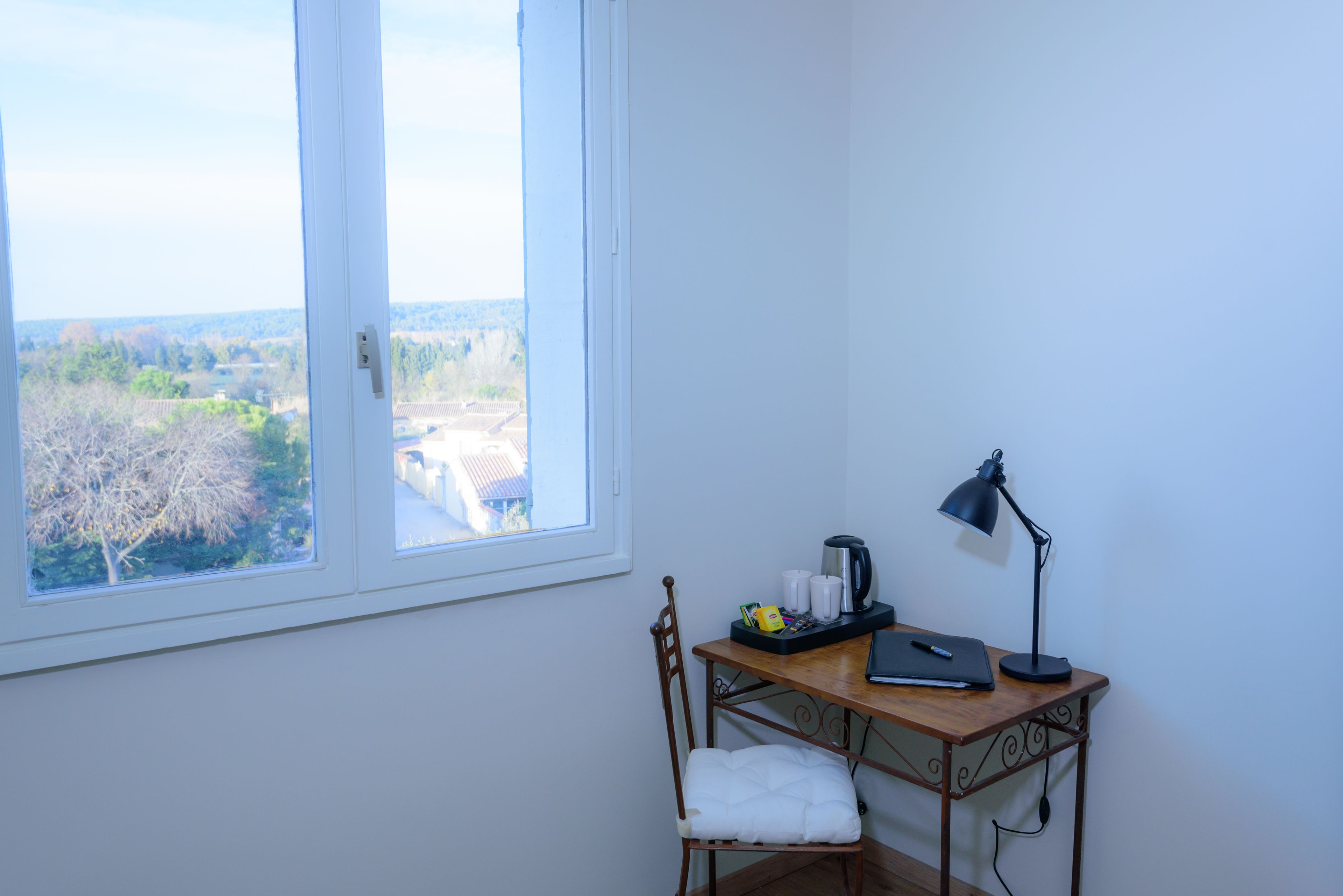 Quarto de canto com escrivaninha, cadeira e vista para uma árvore e prédios.