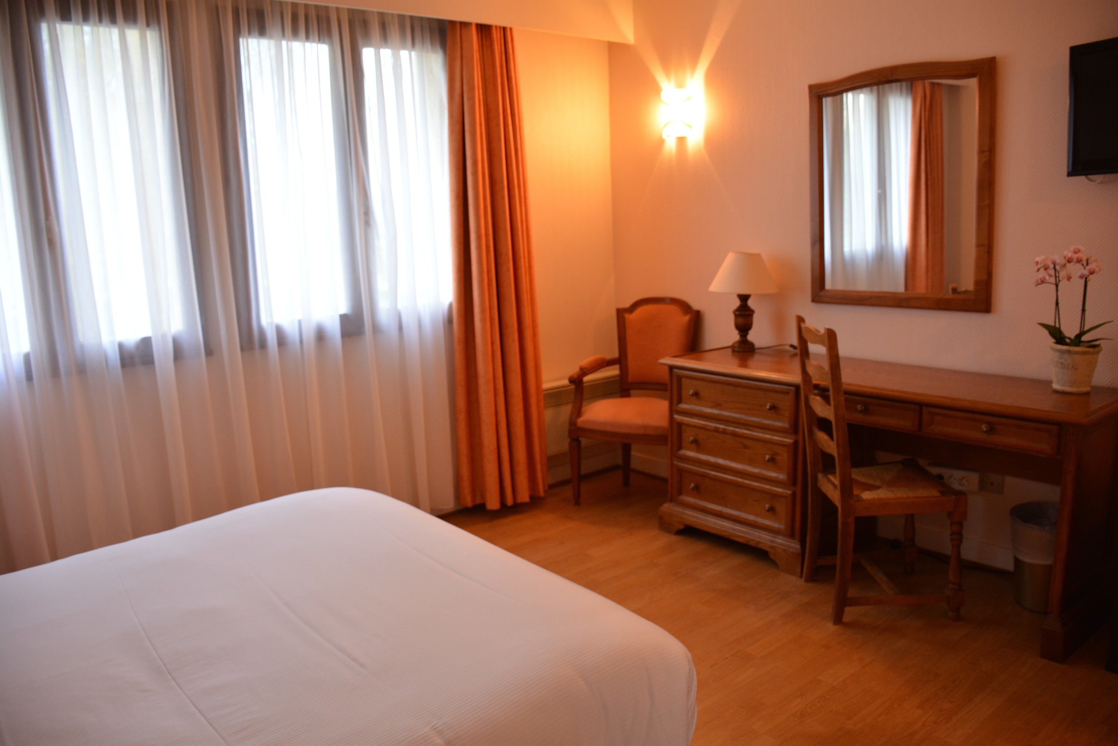 Una habitación de hotel con cama, escritorio con lámpara, silla, cómoda, espejo y ventana con cortinas.