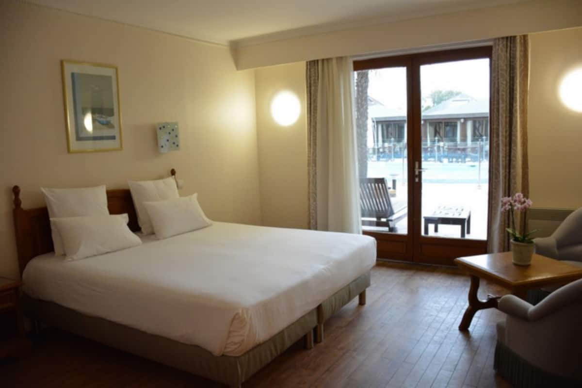 Ein Hotelzimmer mit einem großen Bett, einem Sessel, einem kleinen Tisch mit einer Vase und Blick ins Freie durch eine Schiebetür.