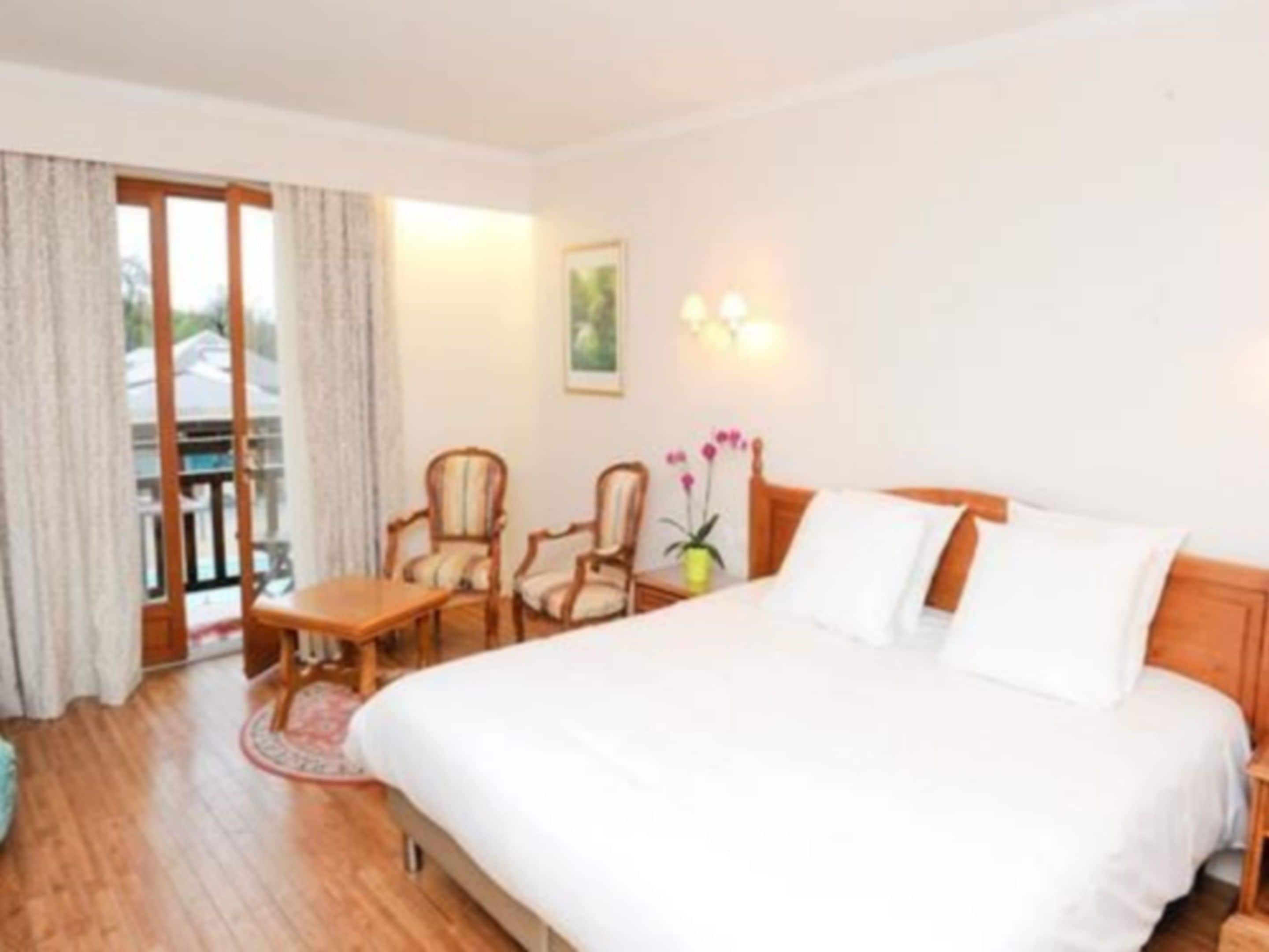 A hotel room with a large bed, two armchairs, a small table, and a balcony with curtains.