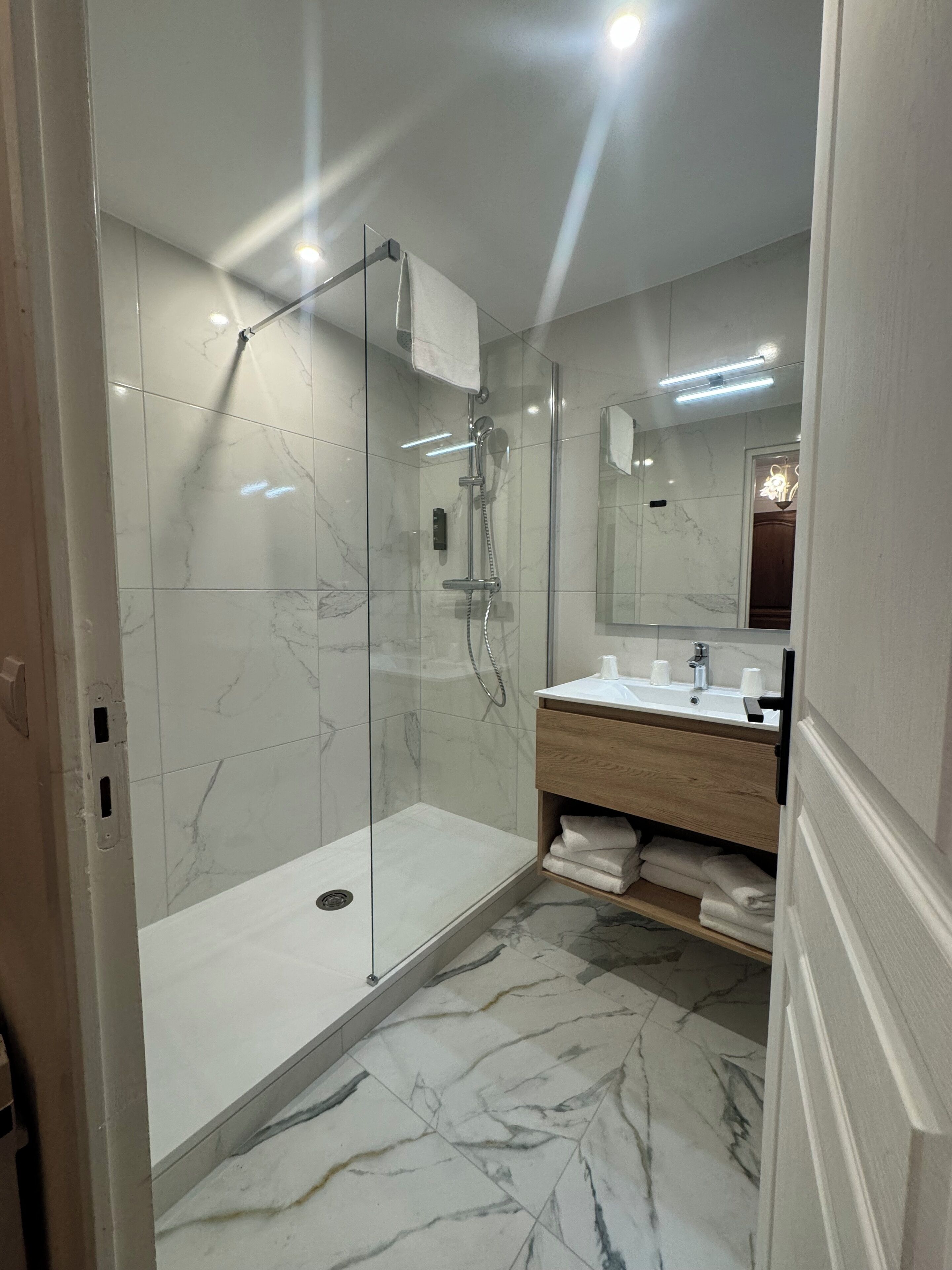 Un baño moderno con ducha envuelta en vidrio, suelo de mármol, tocador de madera y un espejo grande.