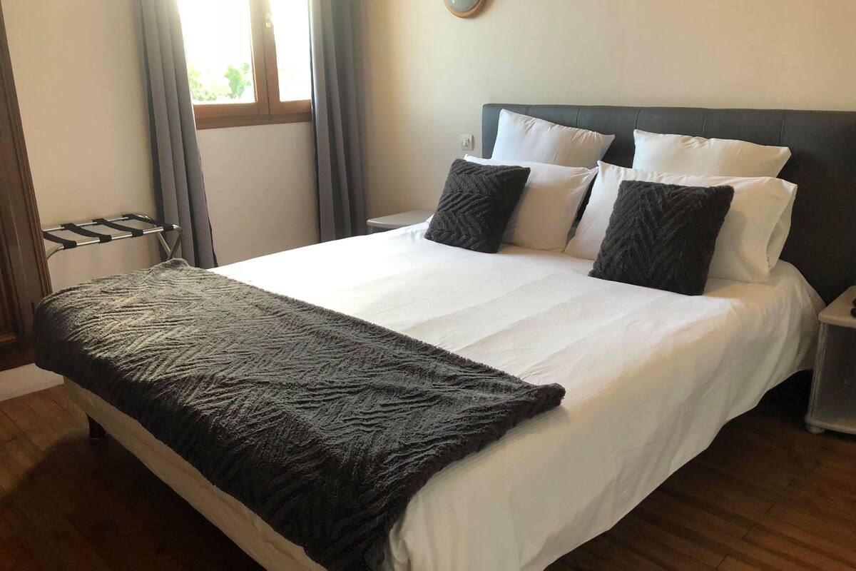 Ein ordentlich bezogenes Bett mit weißer und dunkelgrauer Bettwäsche, ein Holzboden und ein Fenster mit Vorhängen.