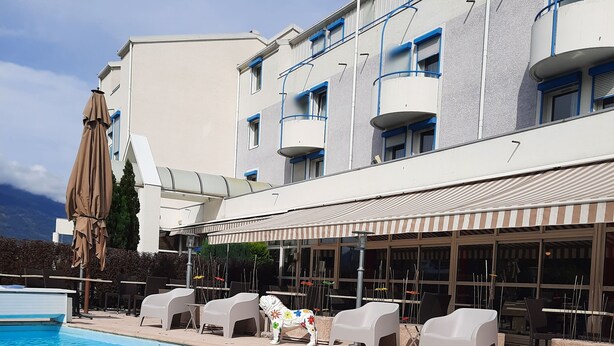 Una piscina con sillones de descanso y sombrillas, un edificio con balcones y vistas a montañas.