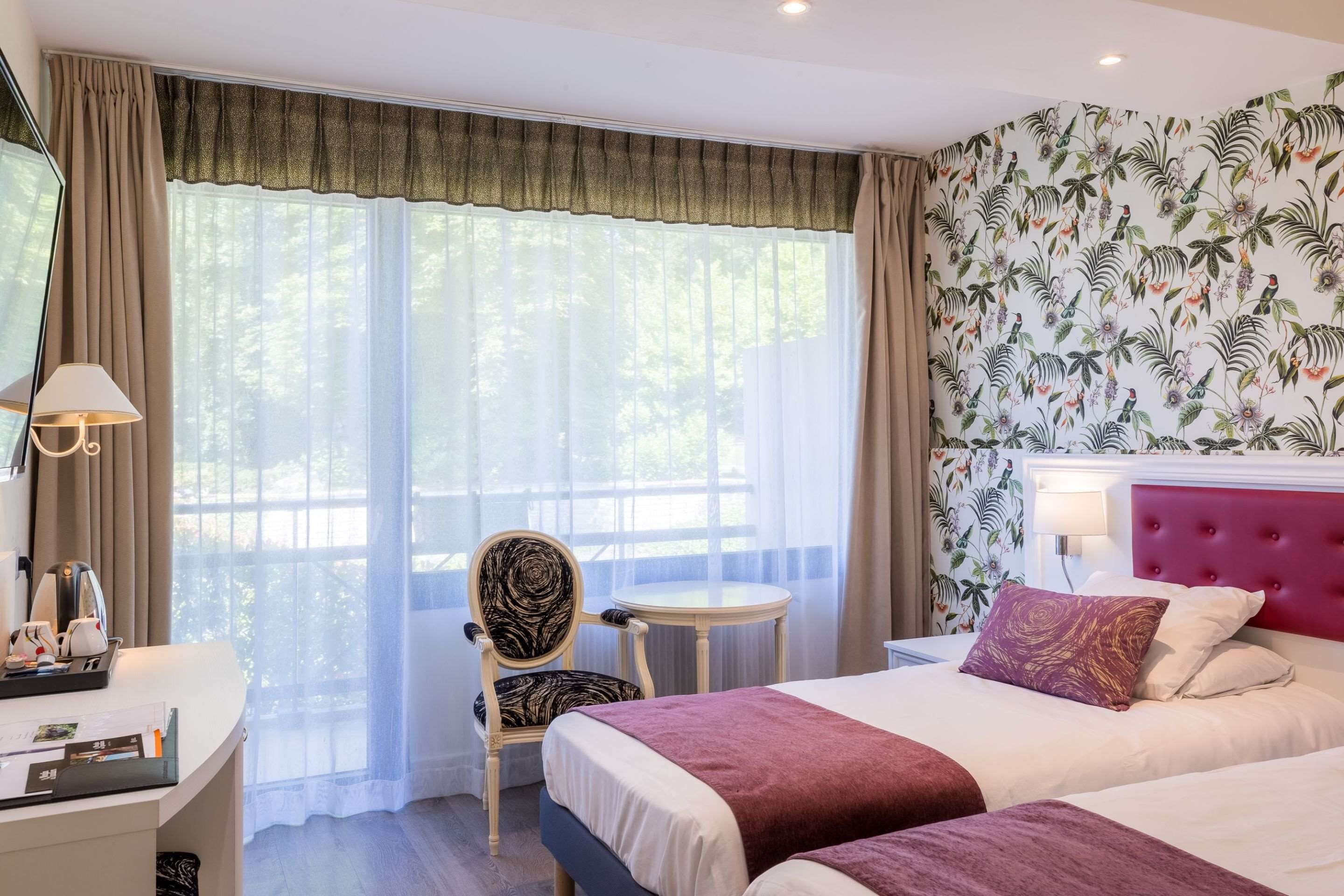 A hotel room with two beds, a desk with a lamp, a chair, and a view of trees through the window.