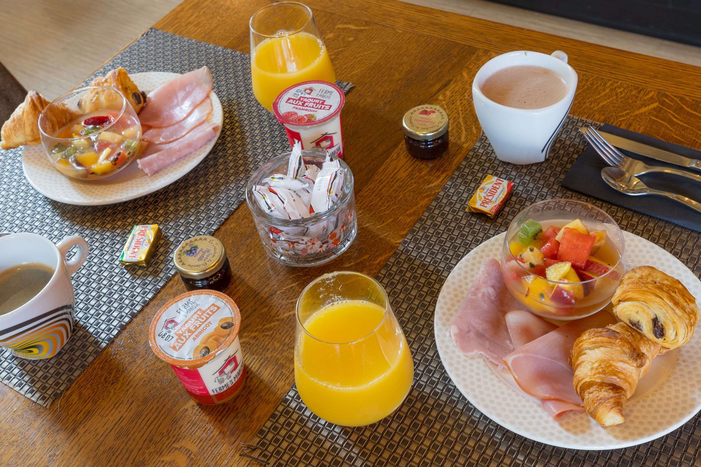 Una mesa de desayuno con cruasanes, jamón, fruta, jugo y parfait de yogur.