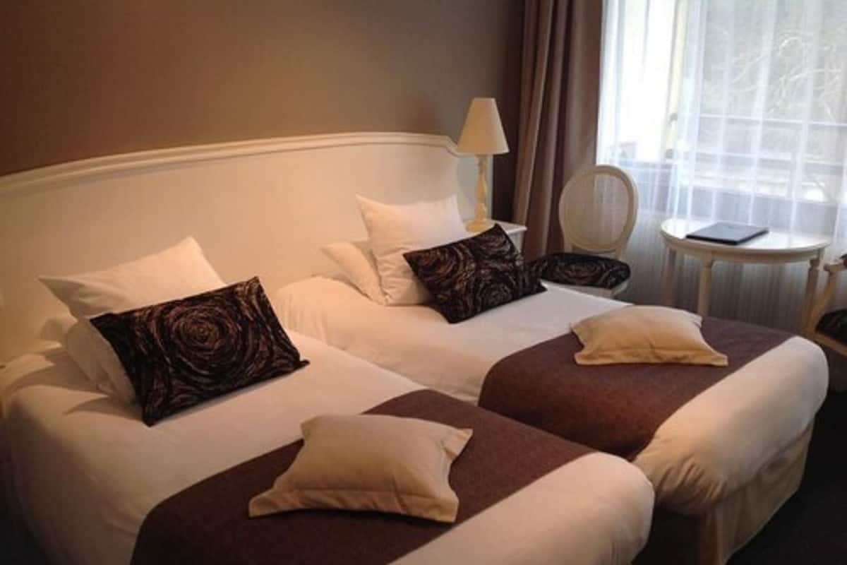 Een hotelkamer met twee bedden, elk voorzien van witte lakens en bruine sierkussens. Er staat een klein rond tafeltje met een lamp en een stoel bij het raam.
