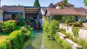 A canal with a water mill, surrounded by lush greenery and traditional buildings with exposed wooden beams and tiled roofs.