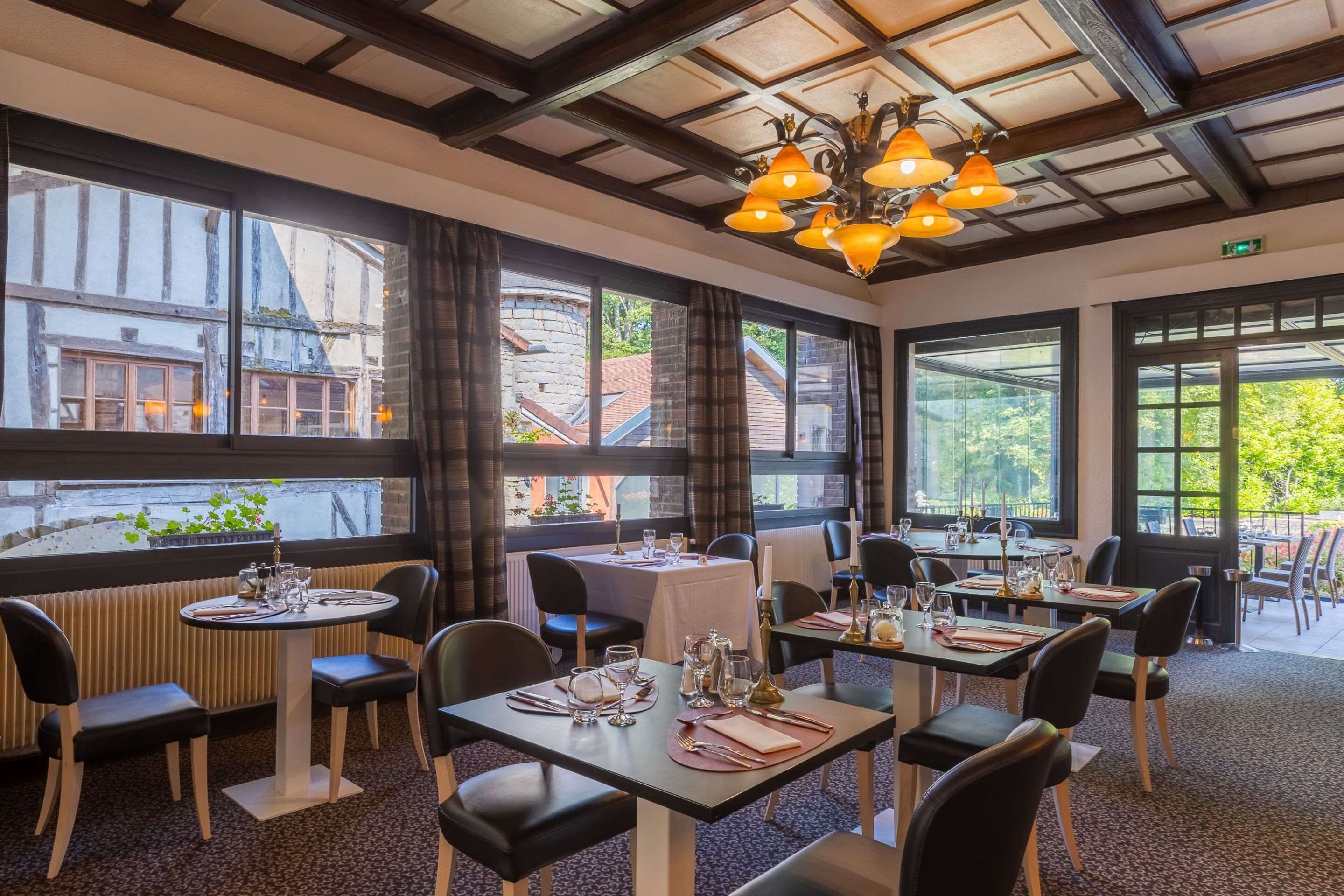 Ein Restaurant mit mehreren gedeckten Tischen, großen Fenstern mit Blick auf Gebäude und Grünflächen sowie einem modernen Kronleuchter.