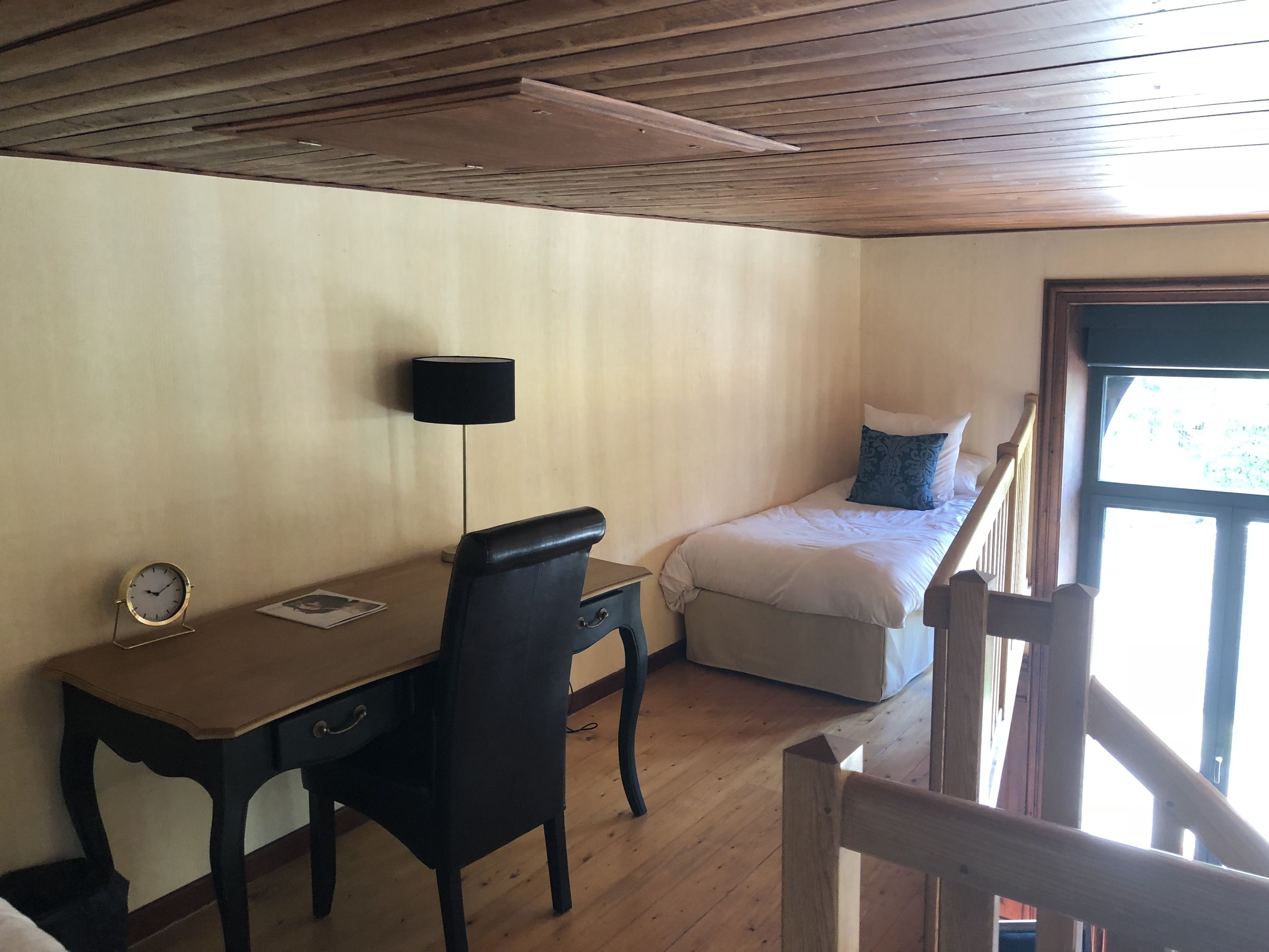 Ein Zimmer mit einem Holztisch, einem Stuhl, einem Bett mit Kissen, einer Lampe und einem Fenster.
