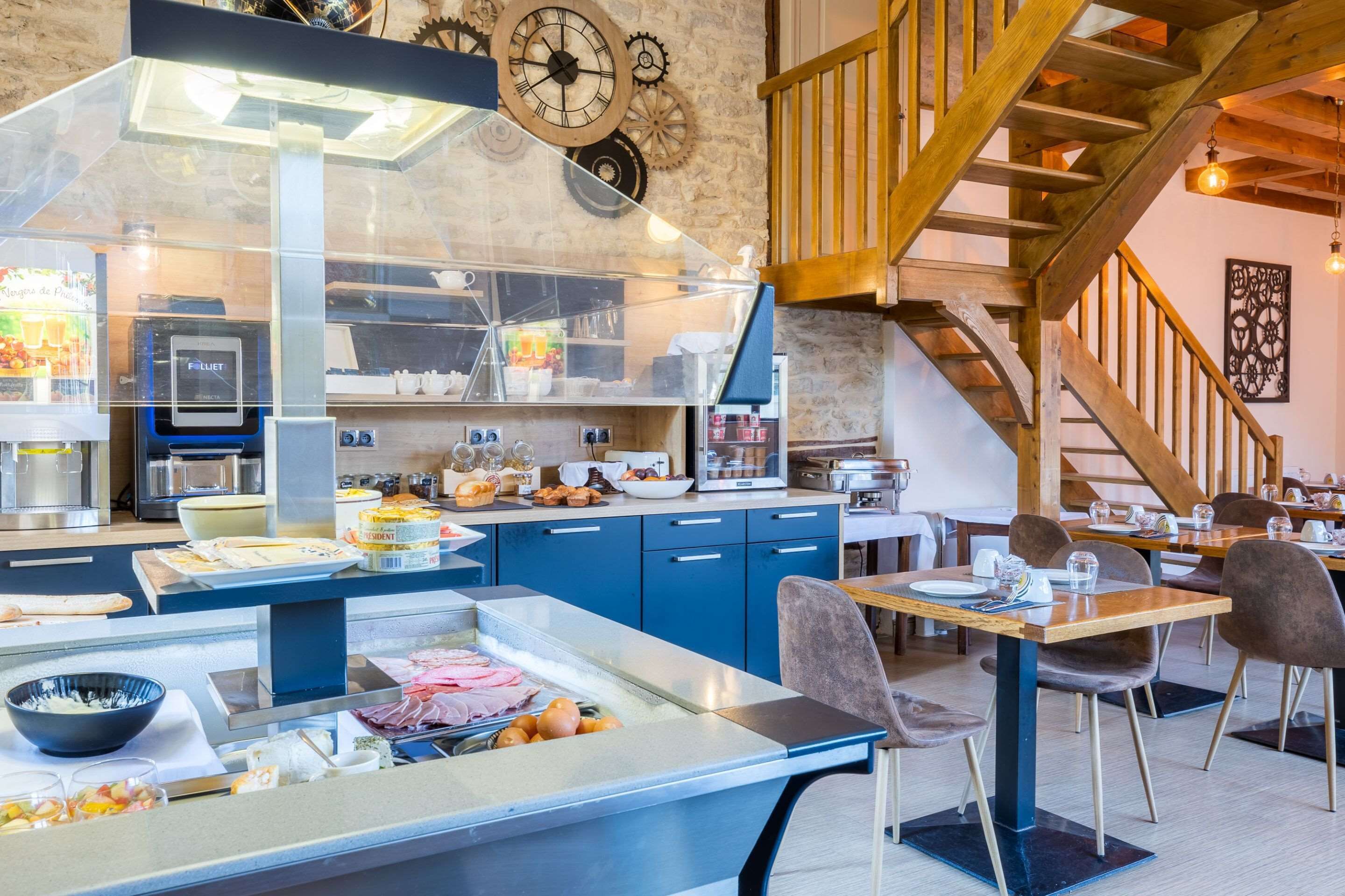 A restaurant with a central counter displaying food, blue cabinets, a wooden staircase, and a clock on the wall.