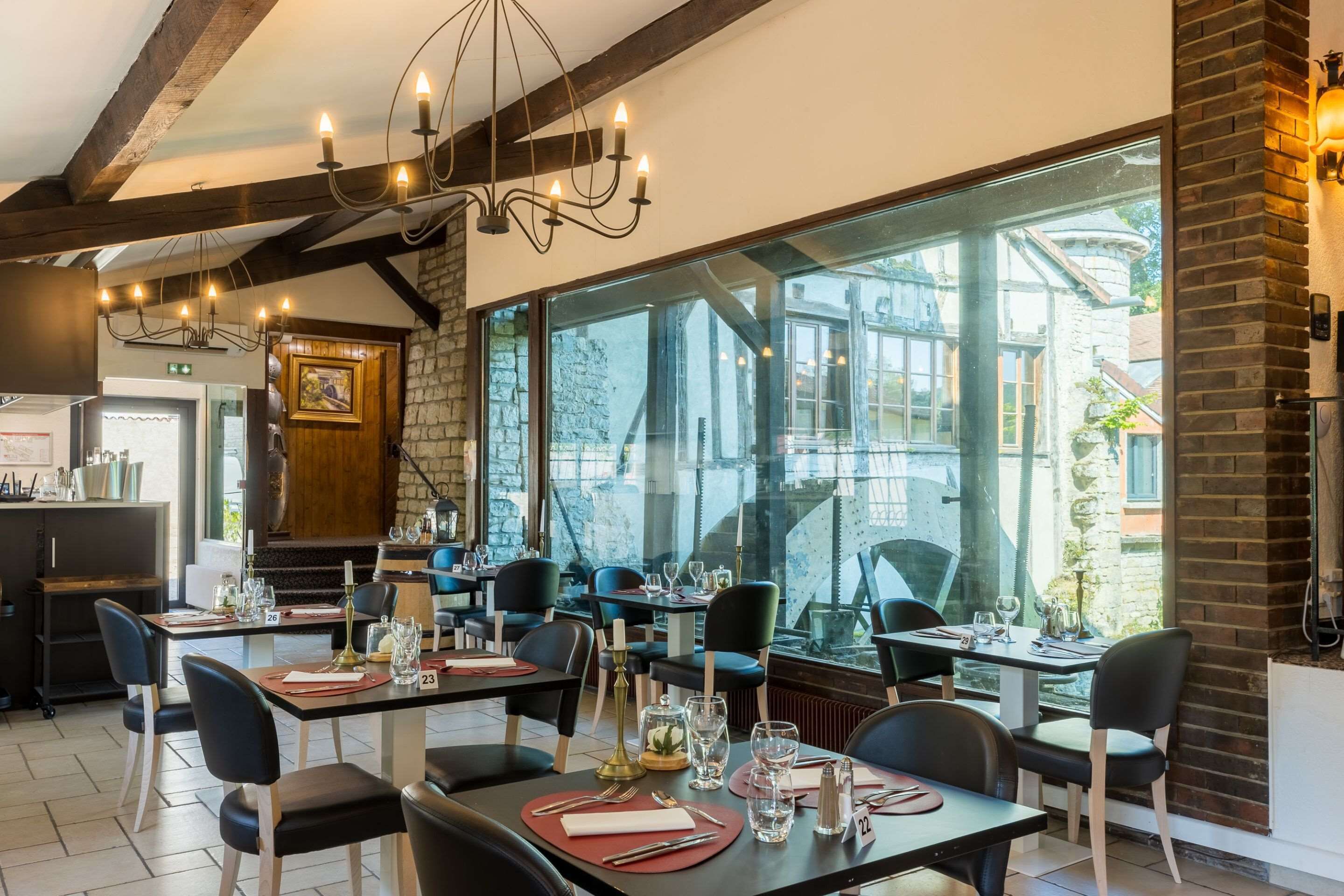 A restaurant with large windows, dark wooden beams, and modern chandeliers. Tables are set with wine glasses and cutlery.