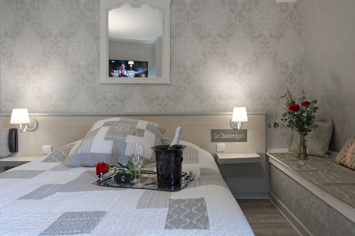 Een slaapkamer met een bed, nachtkastje, spiegel, televisie en een vaas met bloemen.