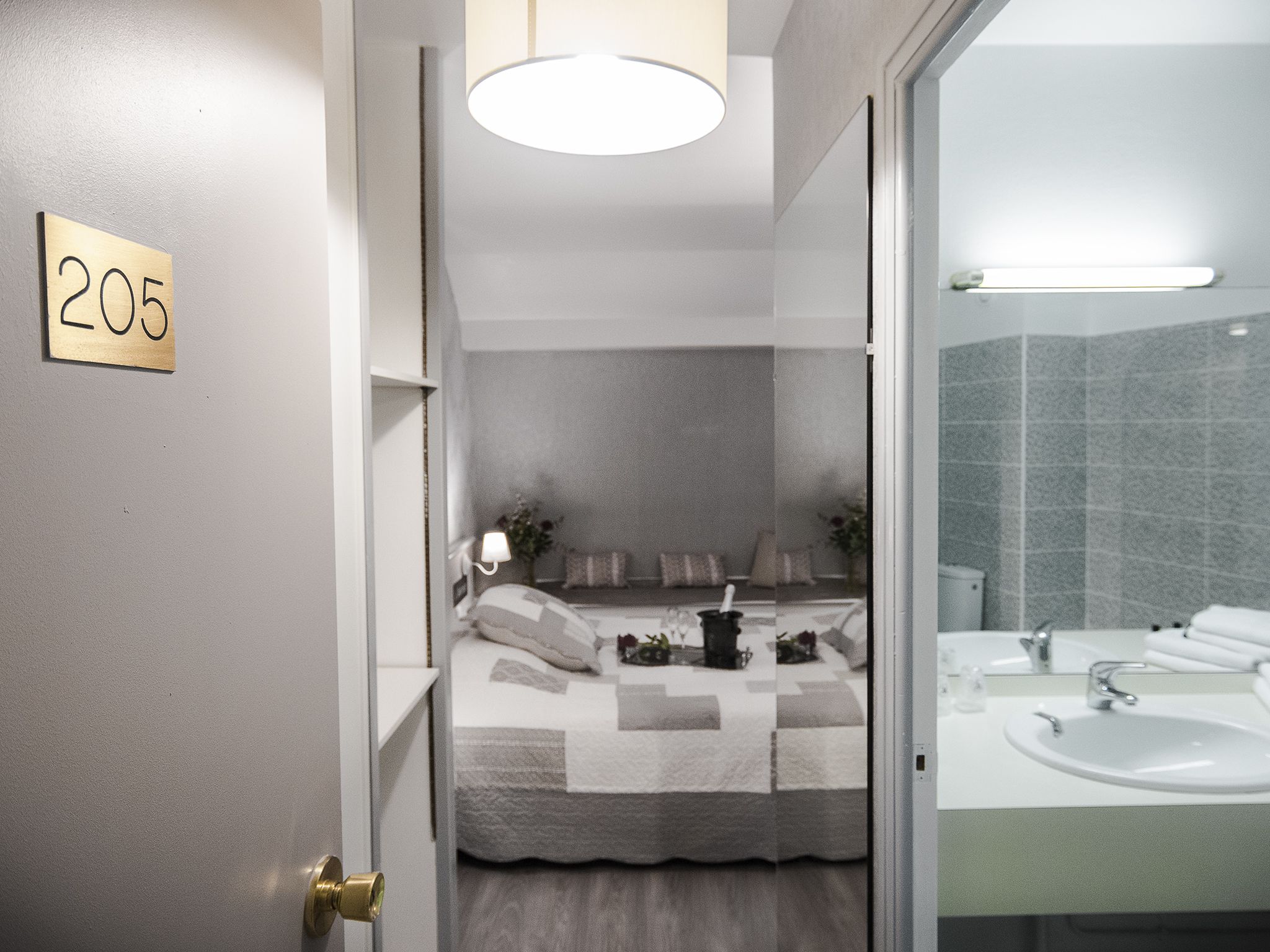 A hotel room with a bed, bedside table, lamp, and a bathroom with a sink and mirror.