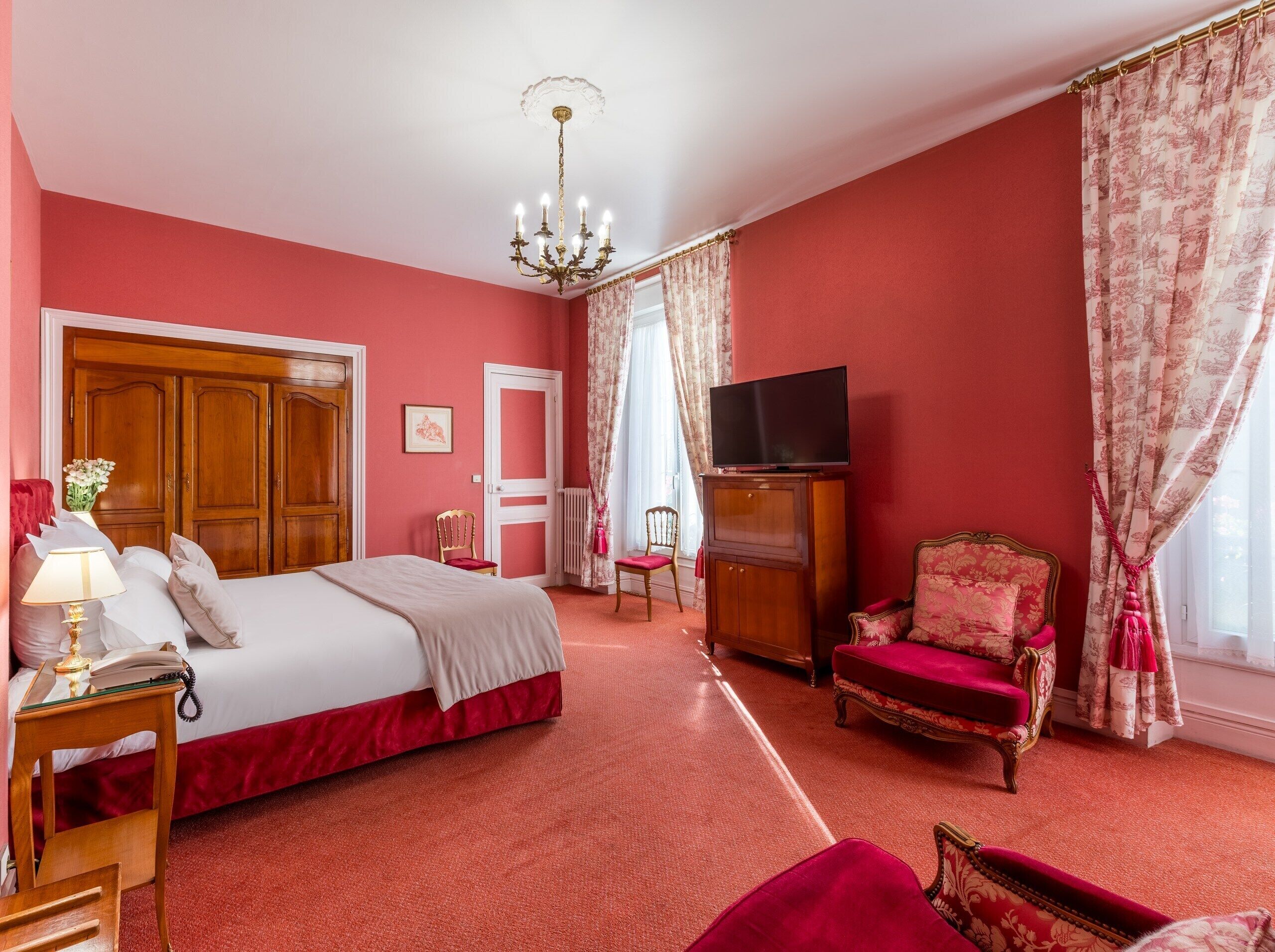 Een hotelkamer met een groot bed, een televisie op een houten dressoir, een rode fauteuil en een kroonluchter.