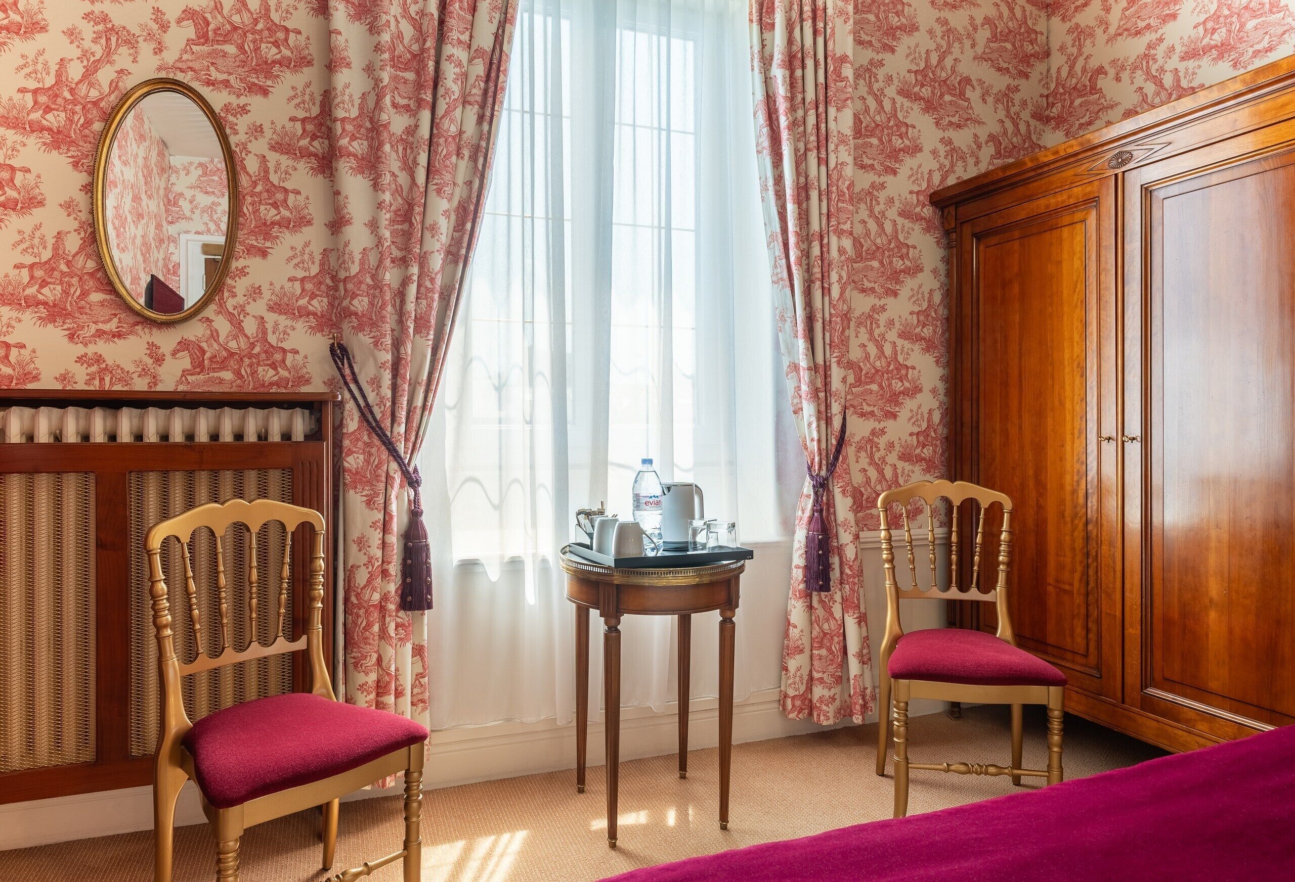 Habitación de hotel con dos sillas de madera, una mesita redonda con una tetera, un radiador y un armario.