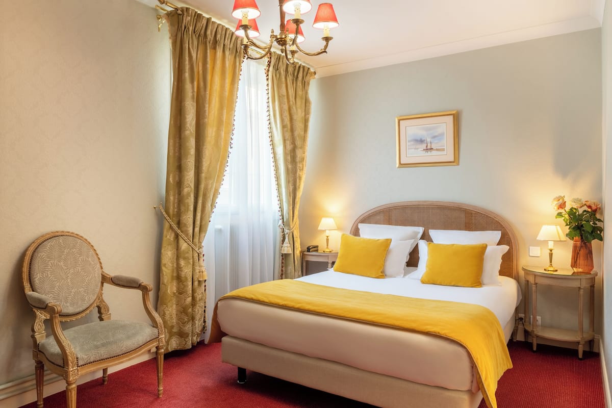 A hotel room with a large bed, a chair, two bedside tables with lamps, a chandelier, and a framed picture on the wall.