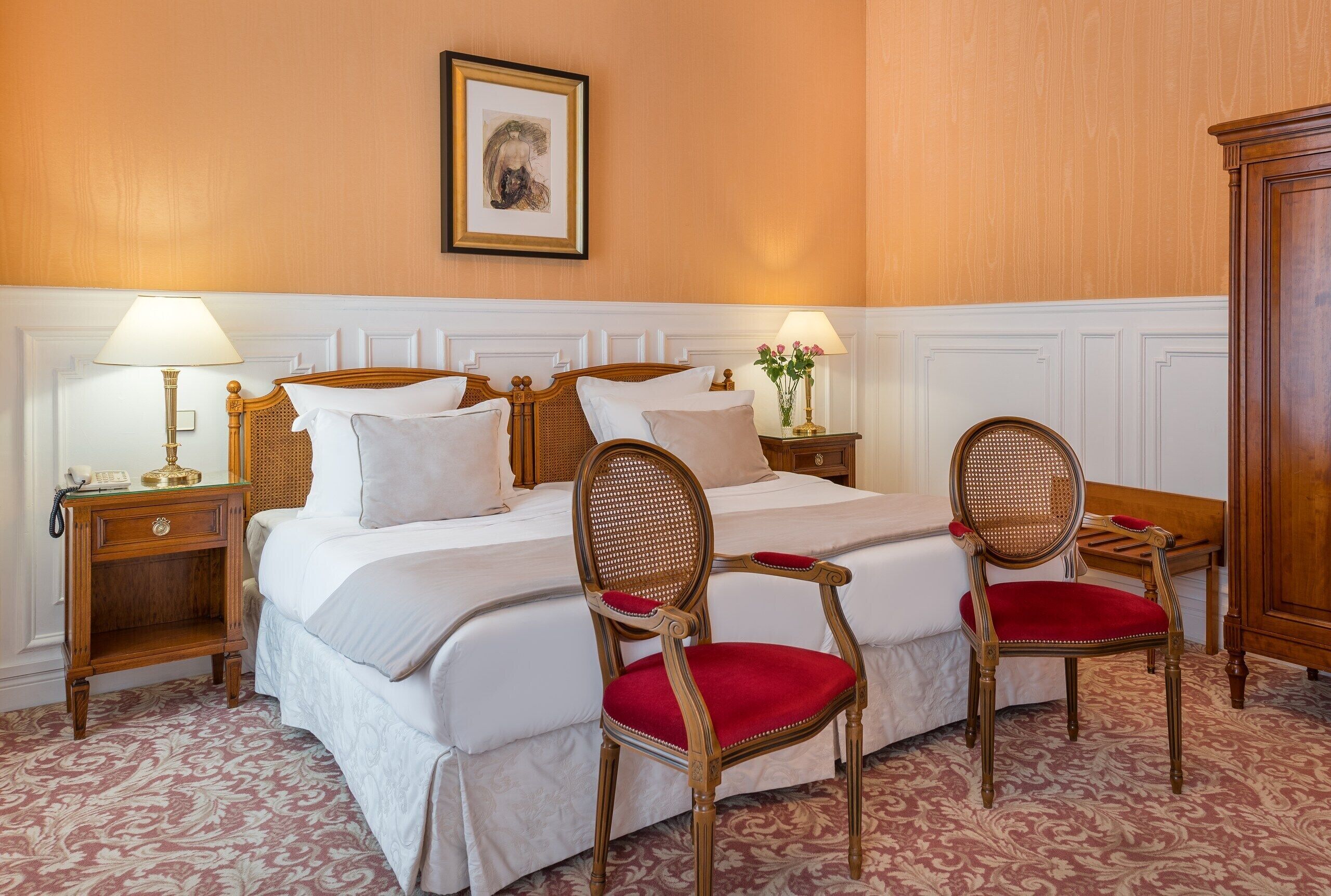 Una habitación de hotel con una cama grande, dos sillas de madera con cojines rojos, una mesita de noche con una lámpara y un cuadro enmarcado en la pared.
