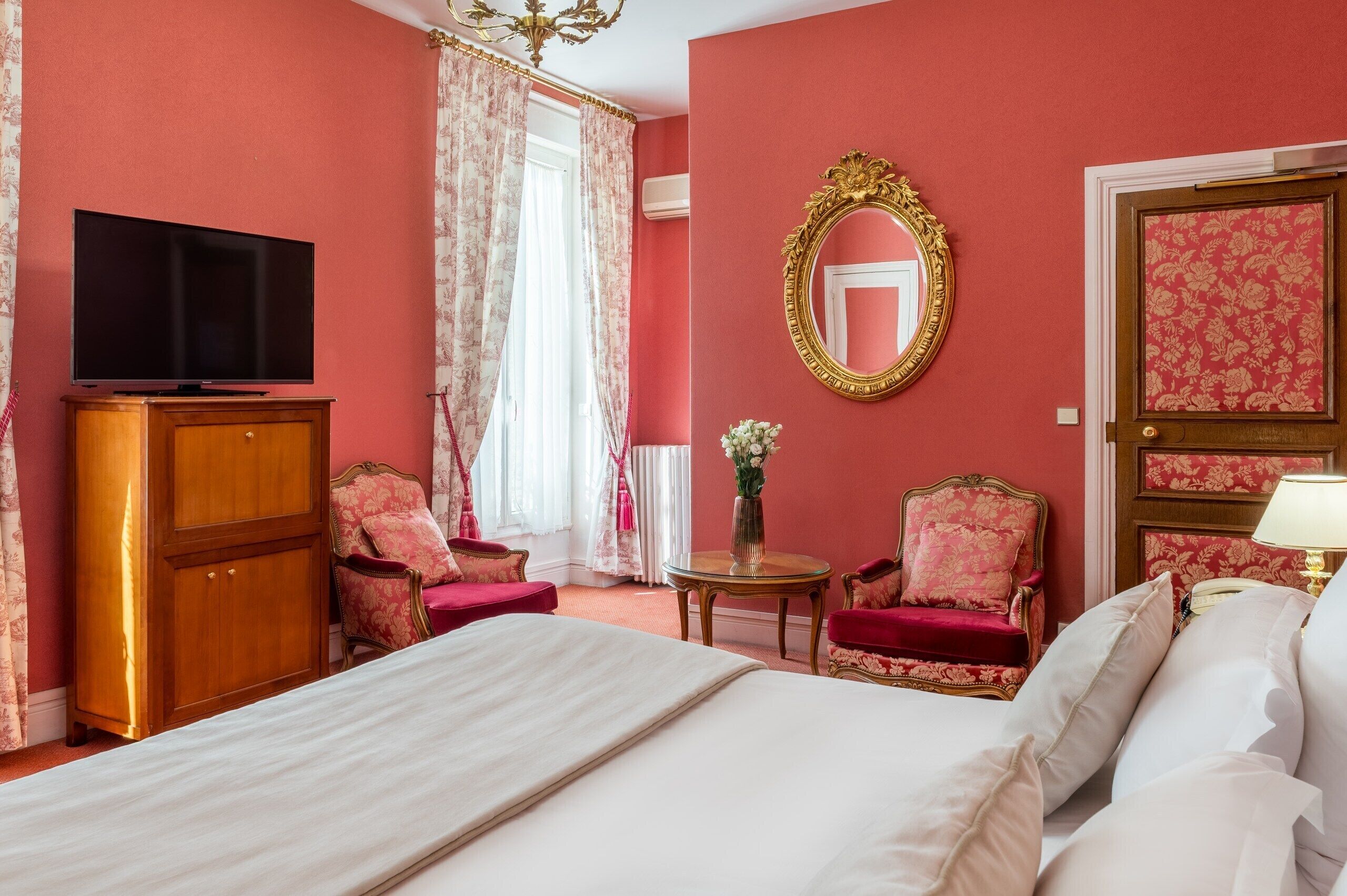 Quarto de hotel com uma cama grande, duas poltronas, uma televisão, um armário de madeira, um espelho e uma porta com papel de parede floral.