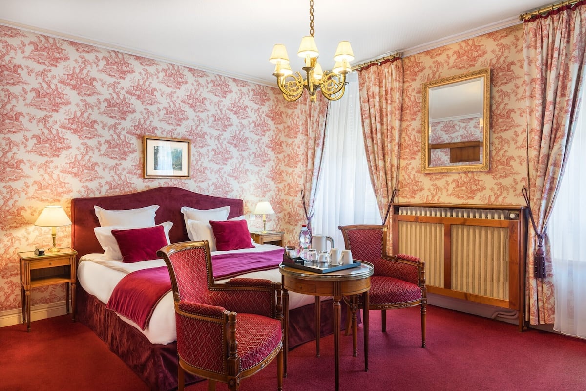A hotel room with a large bed, two armchairs, a small table, a radiator, and a chandelier.