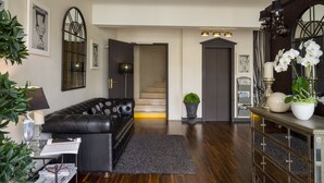 A modern living room with a leather sofa, a coffee table, and a potted plant. There is a staircase leading to another level, a door with a mirror, and a console table with decorative items. - The Originals City, Hôtel Napoléon, La Roche-sur-Yon (La Roche-sur-Yon)