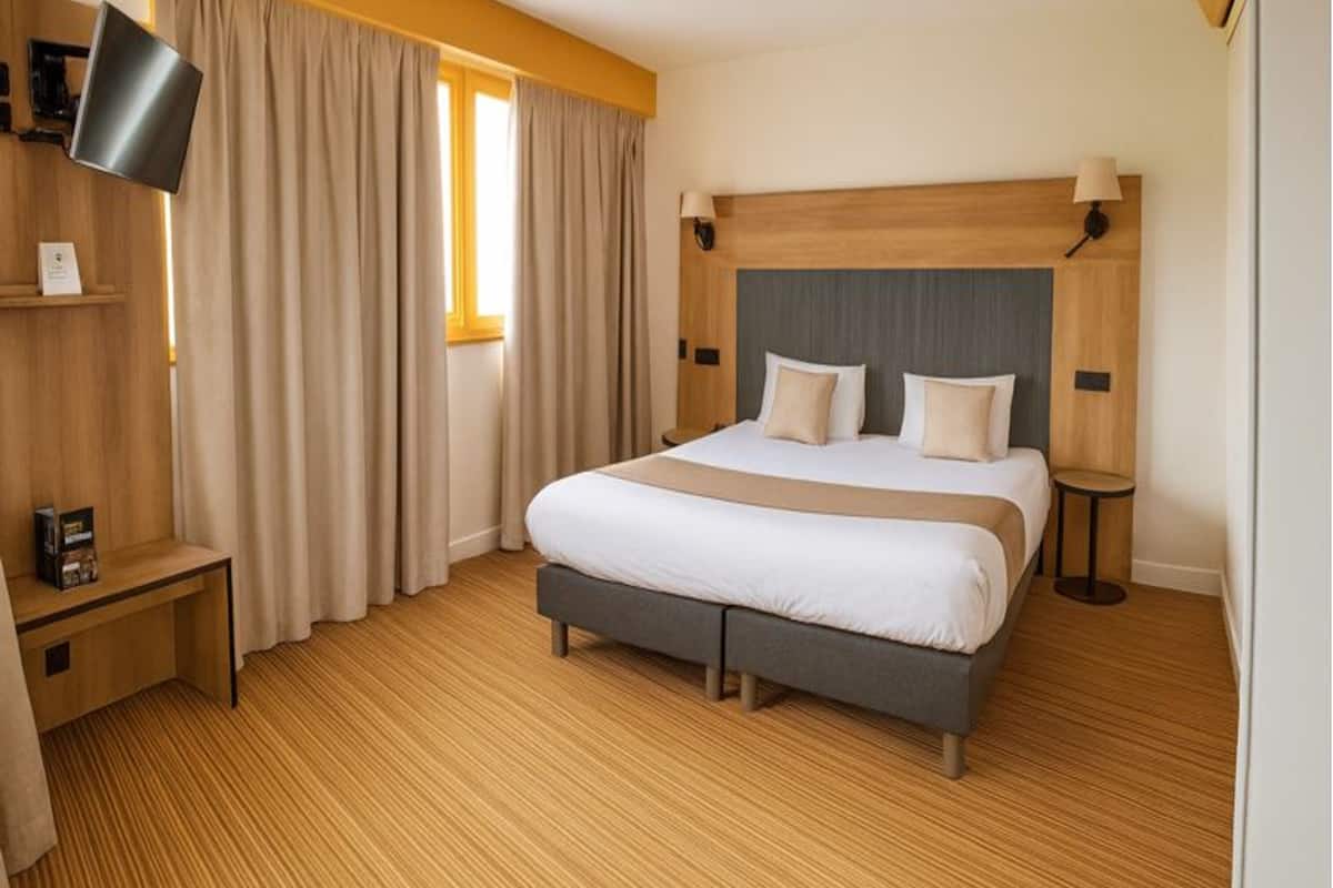 A modern hotel room with a large bed, wooden flooring, a flat-screen TV mounted on the wall, and a bedside table with a lamp.