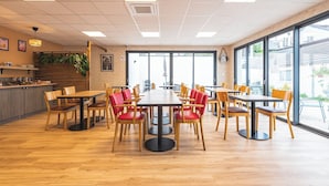 A modern dining area with wooden tables and chairs, a kitchen counter, and large windows offering a view of the outside. - Logis Hôtel Au Tambour (Reims)