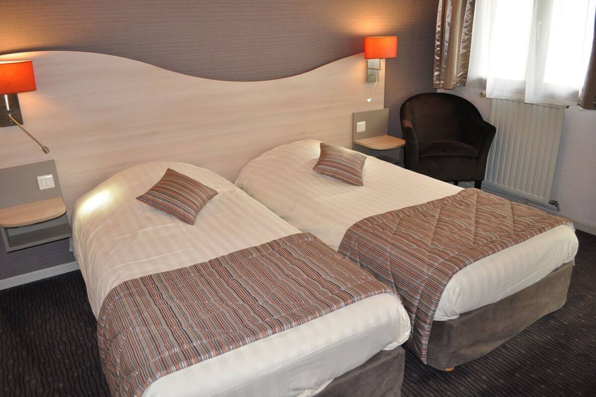 A hotel room with two beds, a nightstand, a chair, and a lamp.