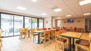 A spacious indoor dining area with wooden tables and chairs, large windows, and a flat-screen TV on the wall.