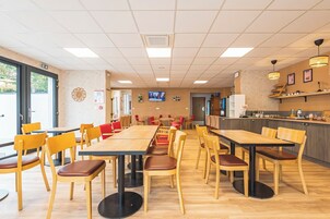 Un'ampia sala da pranzo con diversi tavoli e sedie, un angolo cottura con un bancone e elettrodomestici, e un televisore montato a parete.