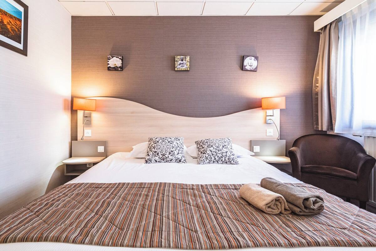 A hotel room with a large bed, two bedside lamps, a chair, and three wall-mounted clocks.